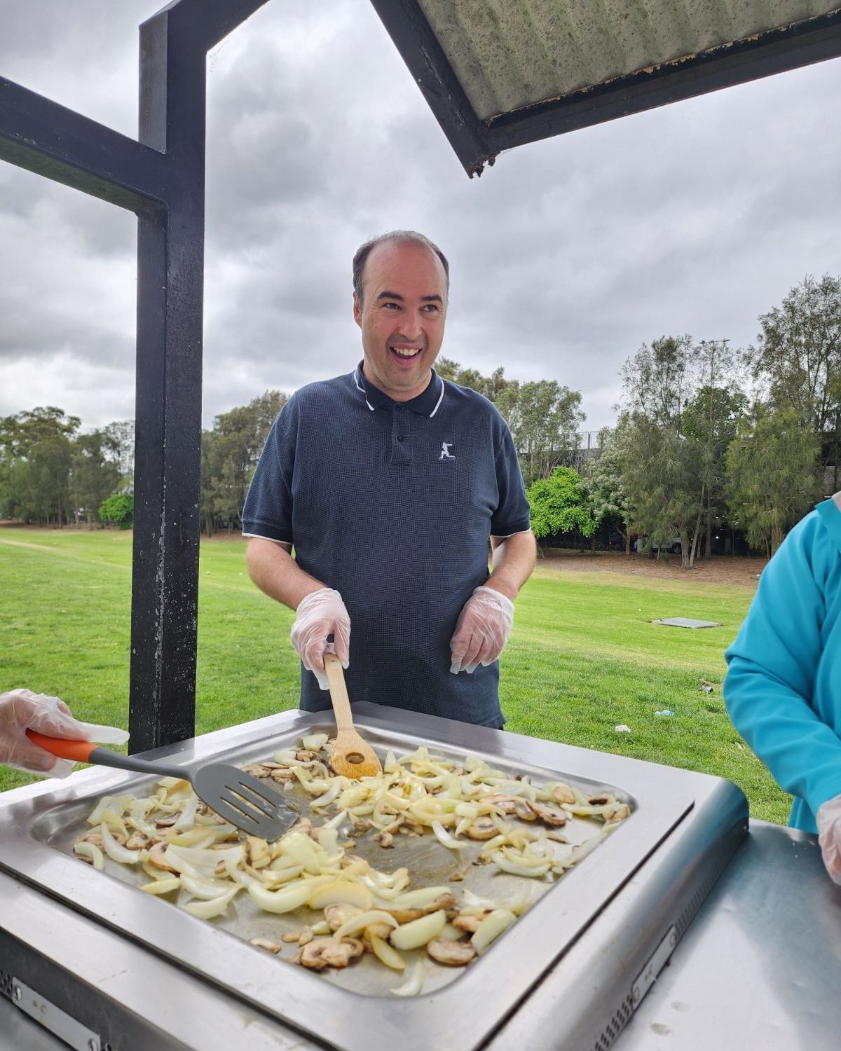 Man cooking onions and mushrooms on a grill outdoors, smiling. Overcast day.