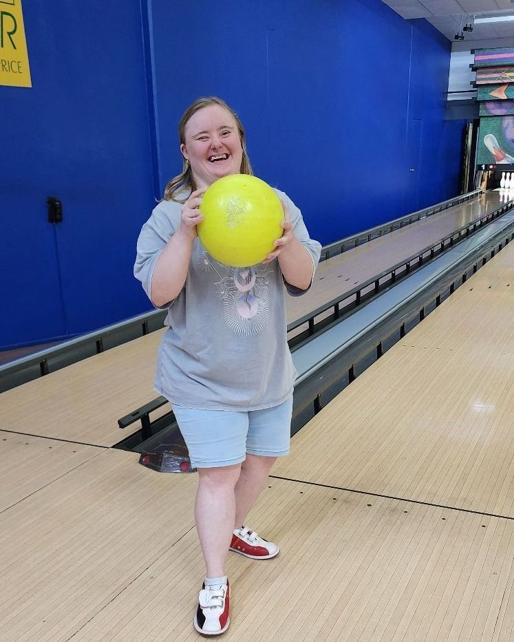 Woman smiling, holding a yellow bowling ball at a bowling alley. She's wearing a gray shirt, denim shorts, and red/white shoes.