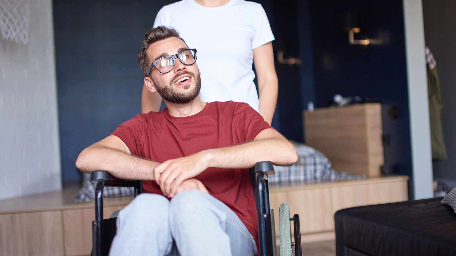 Smiling man in a red shirt sitting in a wheelchair indoors, with another person standing behind him.