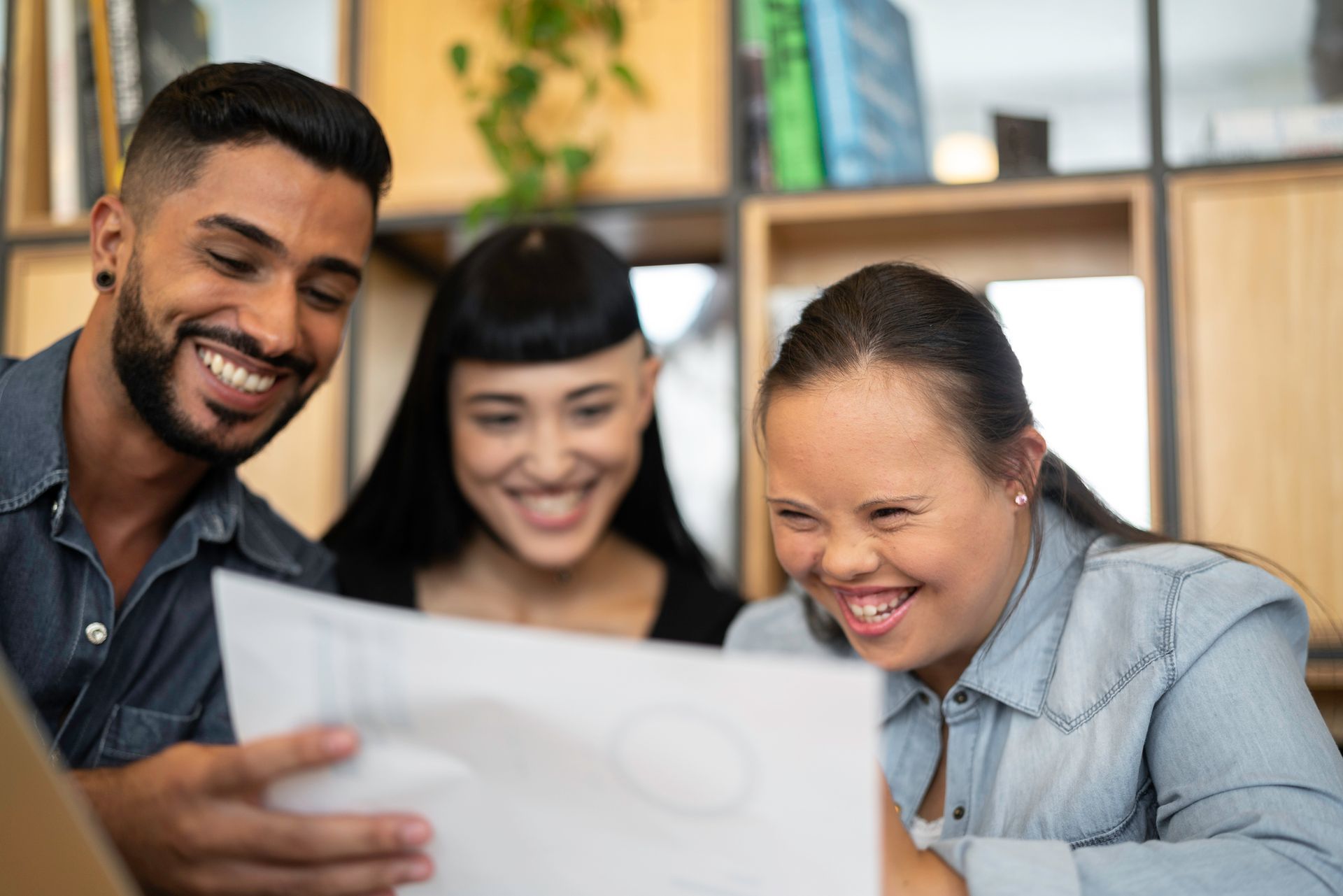Three people smiling, looking at a paper. One woman has Down syndrome. Indoors, near shelves.
