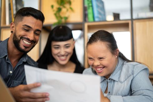 Three people smiling, looking at a paper. One woman has Down syndrome. Indoors, near shelves.