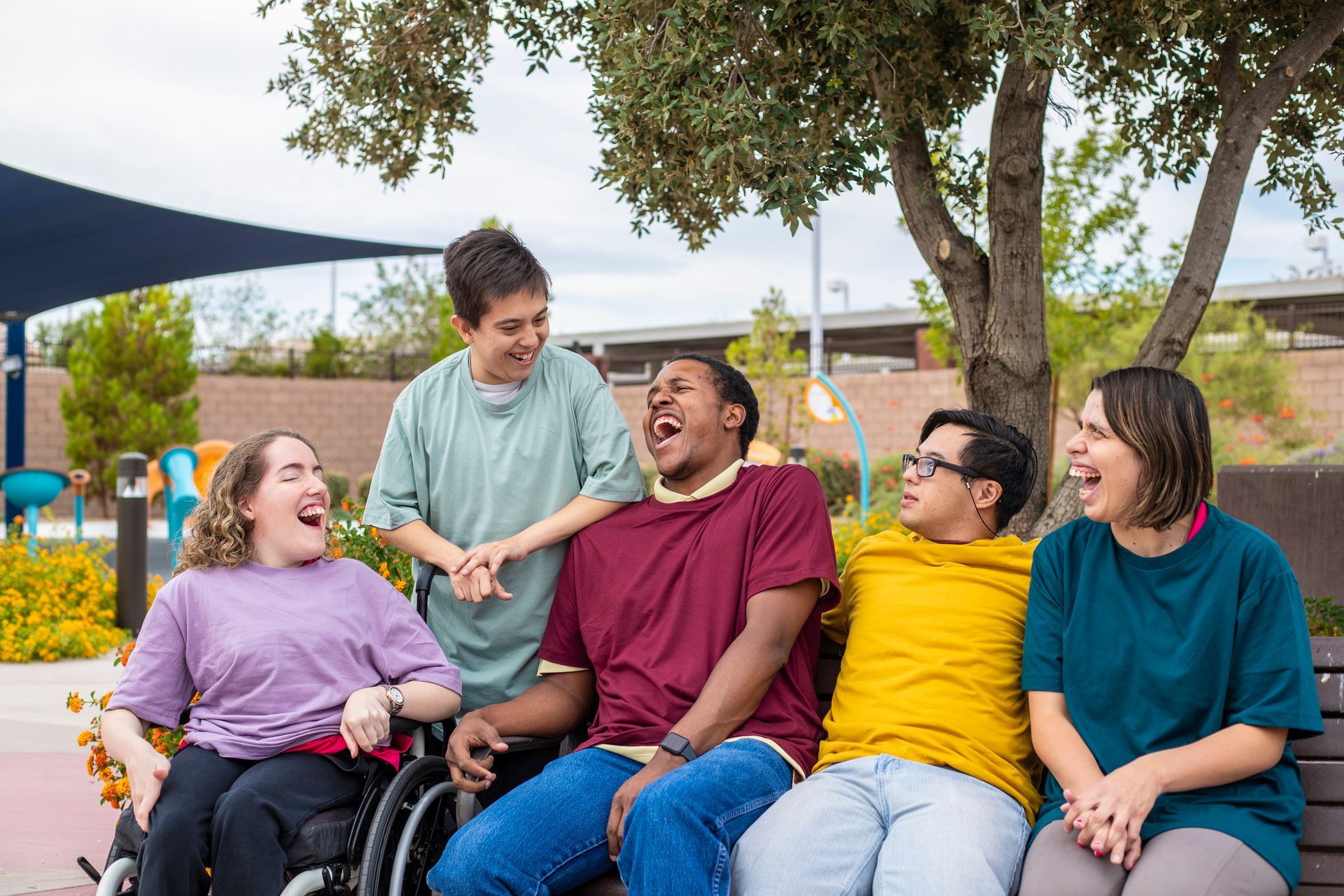 Five smiling people, some in wheelchairs, laugh together on a bench in a park setting.