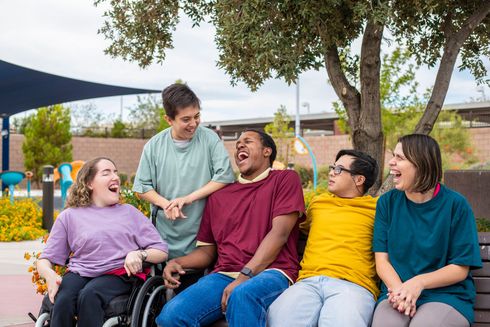 Five smiling people, some in wheelchairs, laugh together on a bench in a park setting.