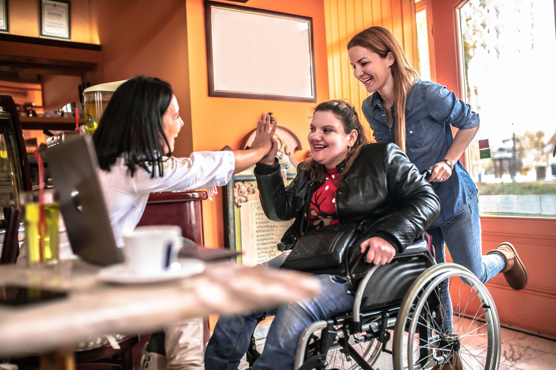 Three people celebrating in a cafe. A woman in a wheelchair high-fives another, smiling, as a third smiles and cheers.