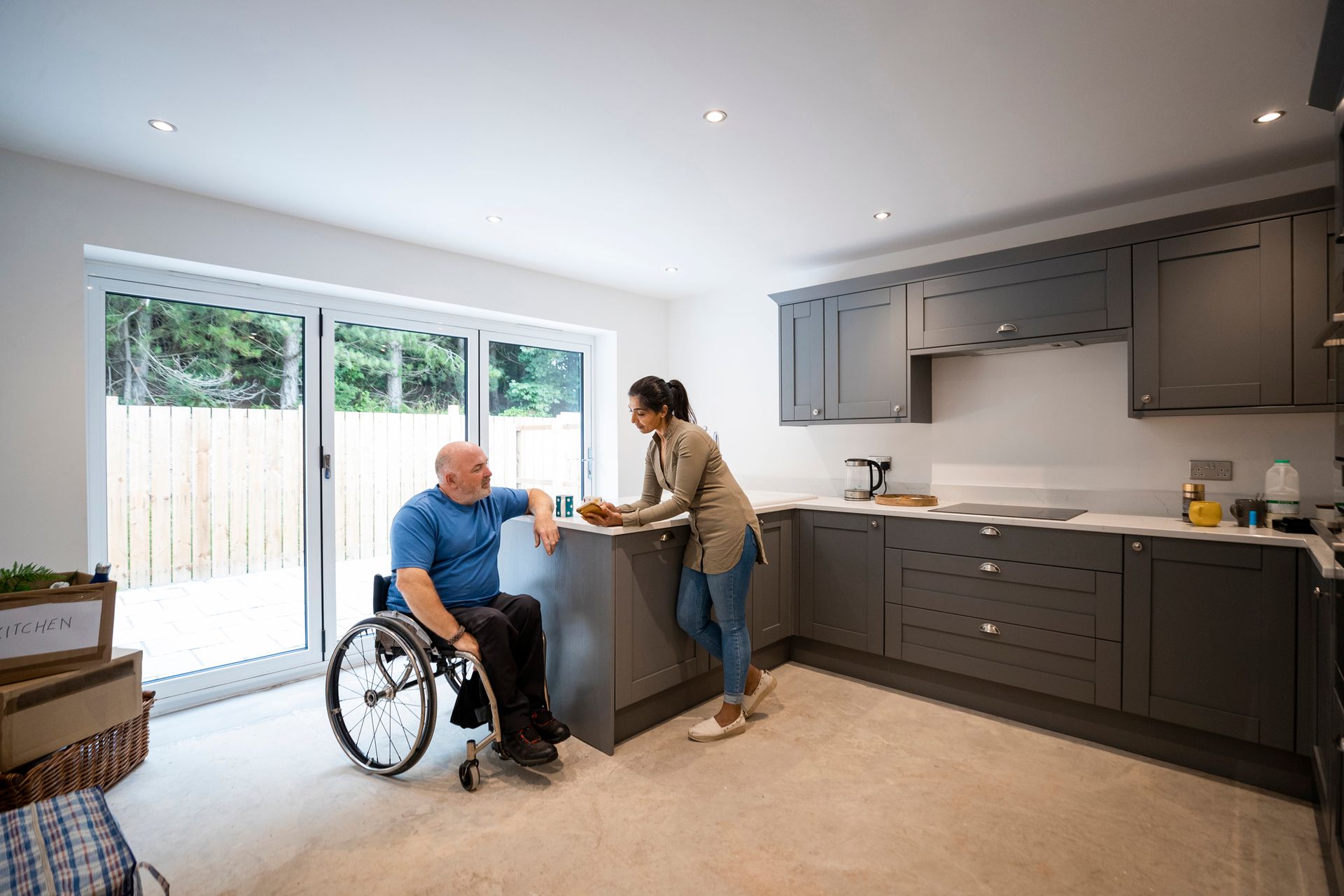 Man in wheelchair and woman in kitchen, discussing; modern setting with gray cabinets.