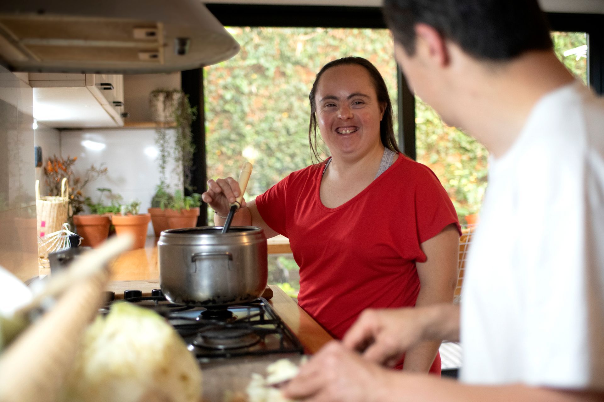 Woman stirs pot on stovetop, smiling. Man in white shirt chops food nearby. Kitchen setting.