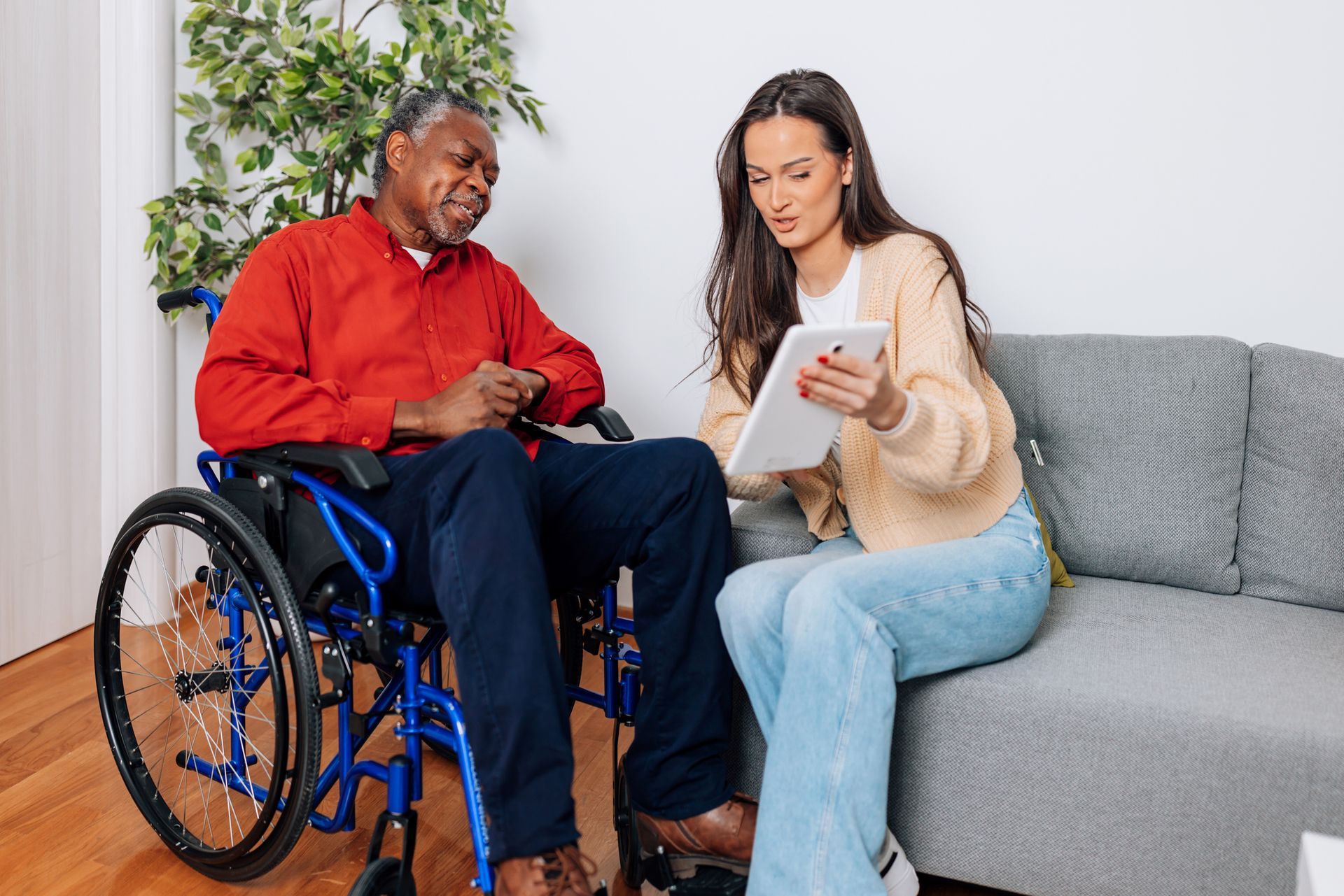 A woman shows a tablet to a man in a wheelchair; both are indoors in a living room setting.