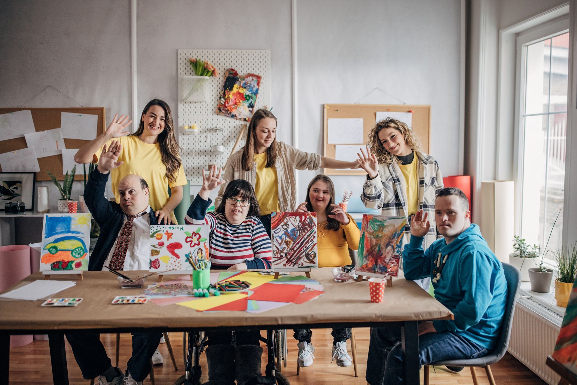People with disabilities and caregivers at an art table, waving and smiling. Studio setting with art supplies and paintings.