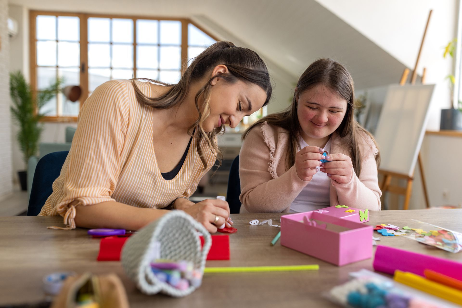Woman and girl smiling, crafting at a table. Sunlight streams in. Pink box and basket with supplies.