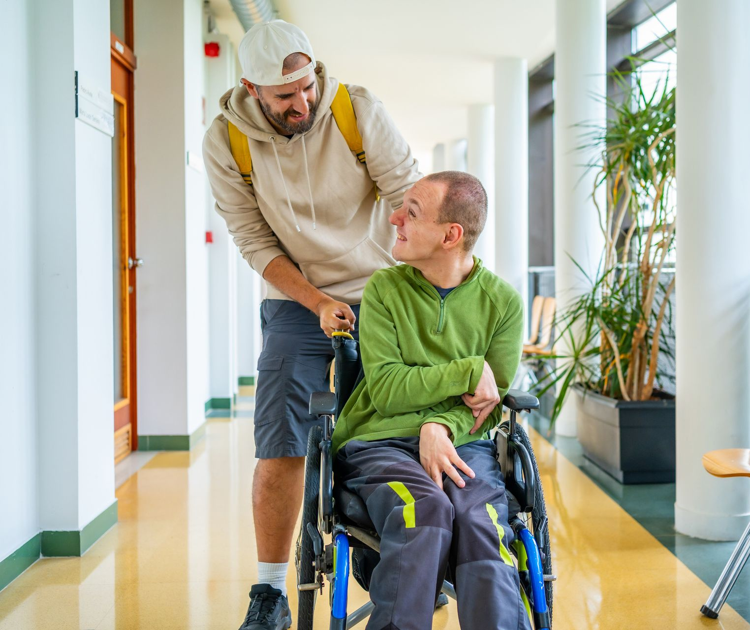 Man in wheelchair being pushed by another man in a hallway. Both men smiling, looking at each other.