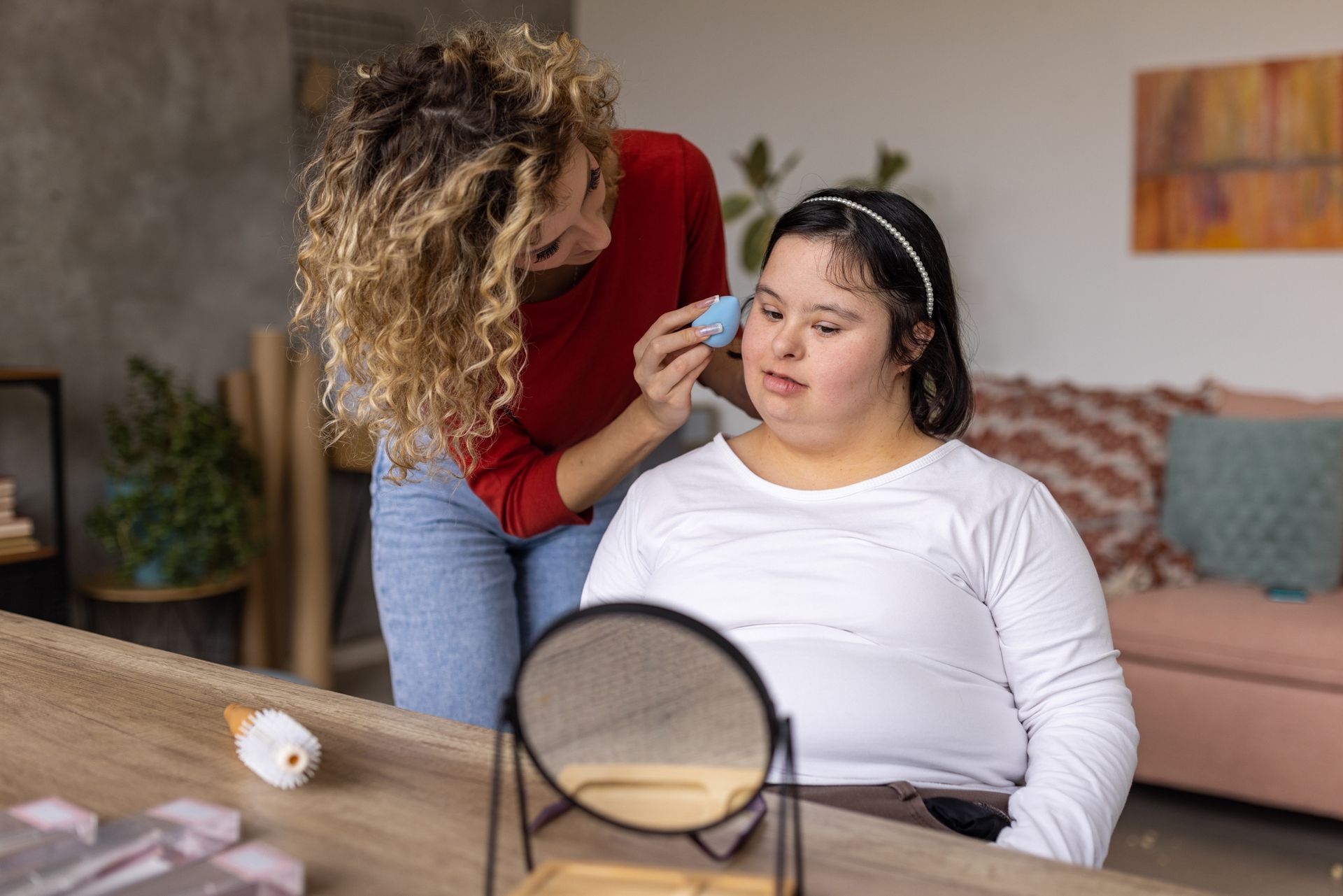 Woman applying makeup to another woman's face, using a sponge, with a mirror on a table indoors.