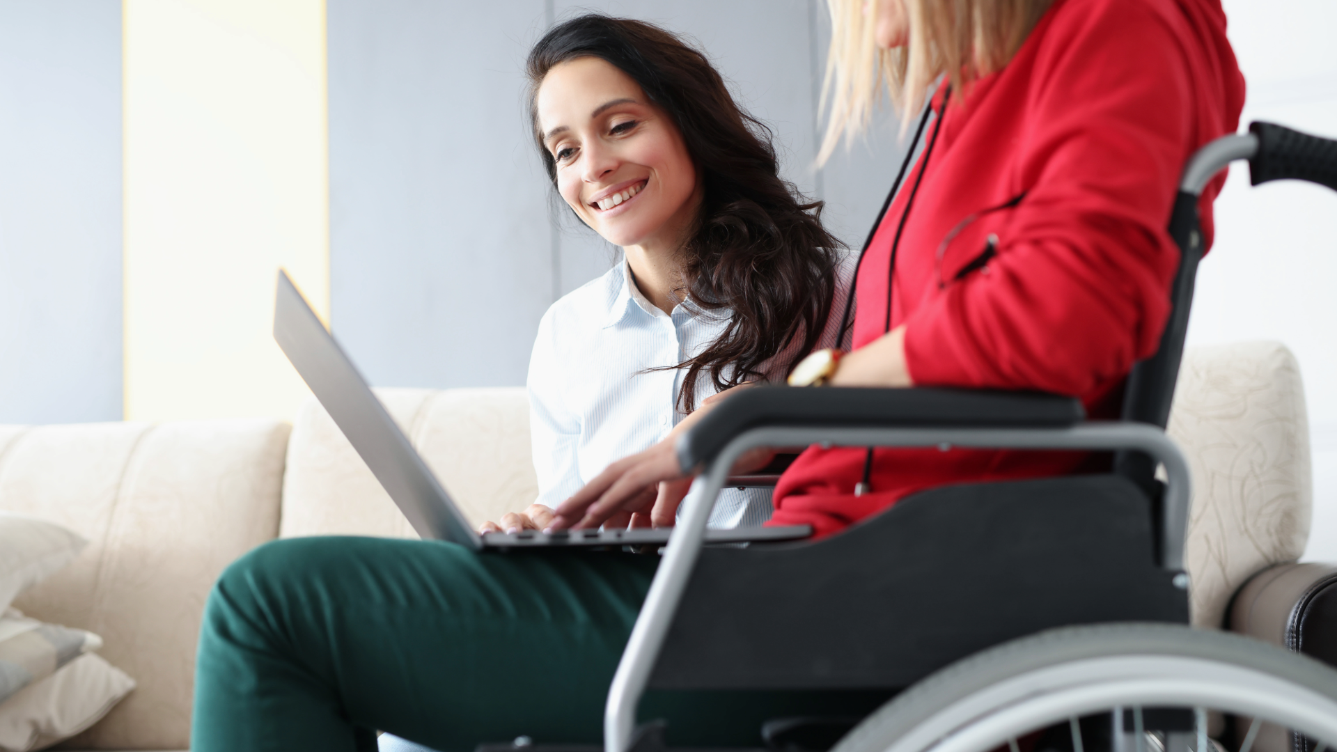 Woman in white shirt smiles beside wheelchair user while looking at a laptop on a couch