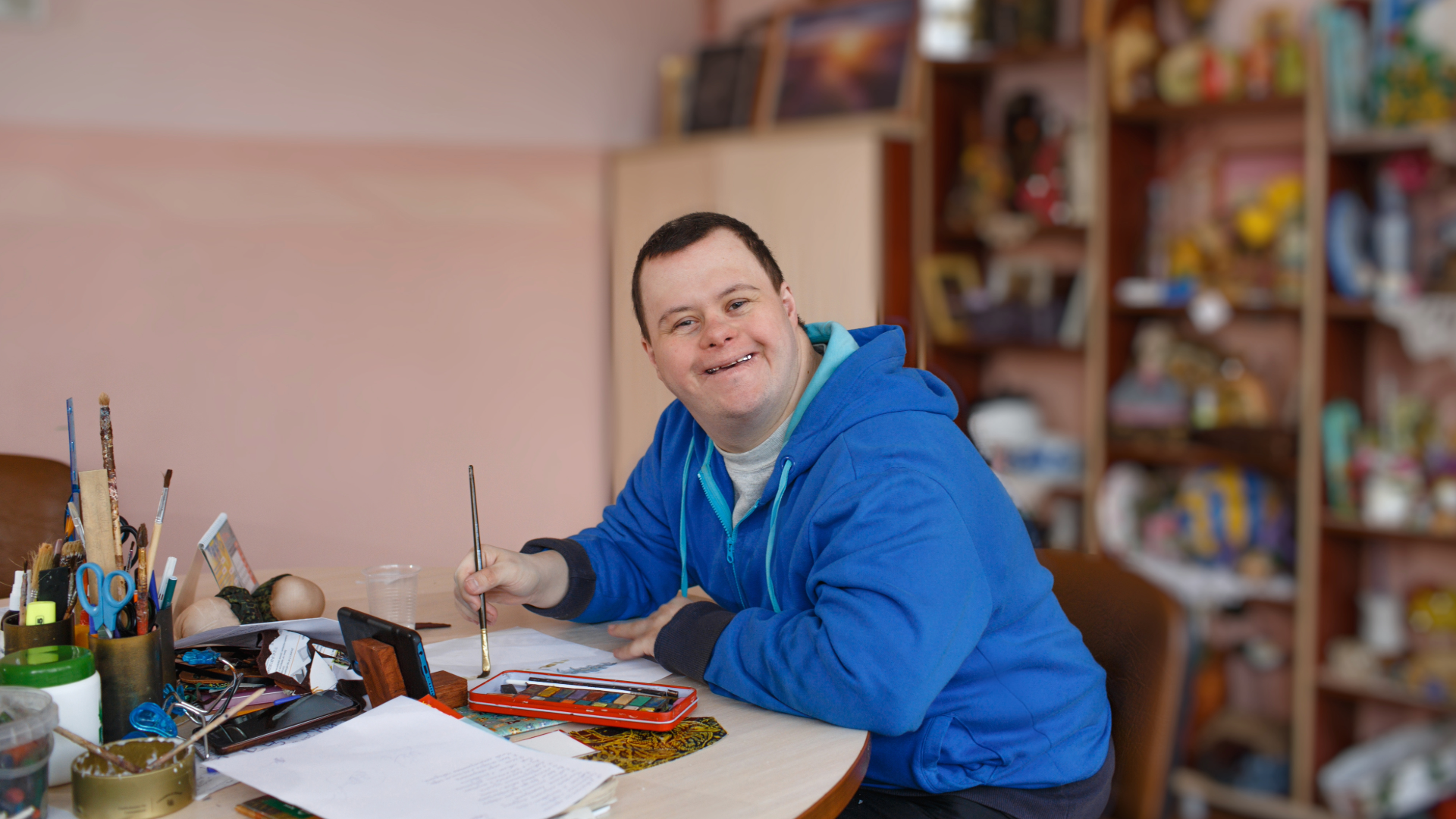 Person in a blue hoodie painting at a desk in a bright studio.