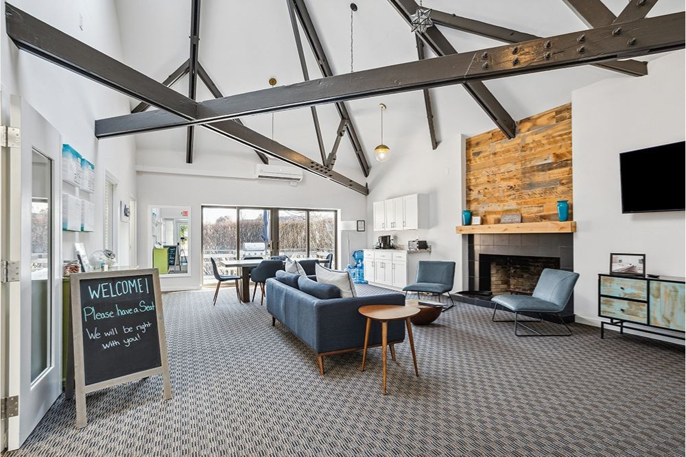 Lounge with a blue sofa, chairs, fireplace, and open ceiling with exposed beams. A welcome sign stands by the door.