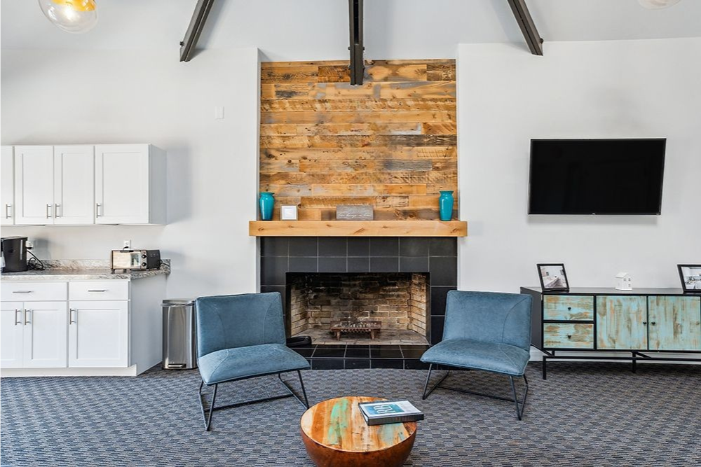 Cozy lounge area with fireplace, chairs, and coffee table. Wood accent wall with logo above fireplace, TV on right.