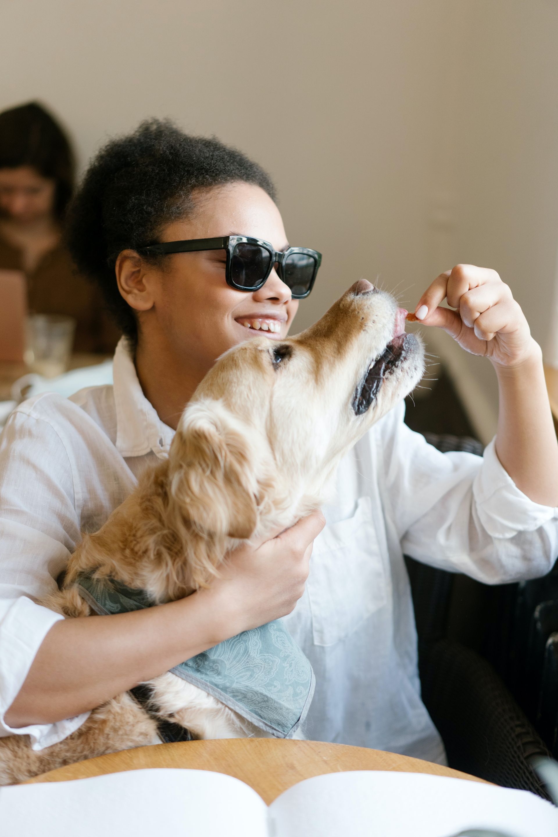 Person in sunglasses feeds treat to a golden retriever, seated in cafe.