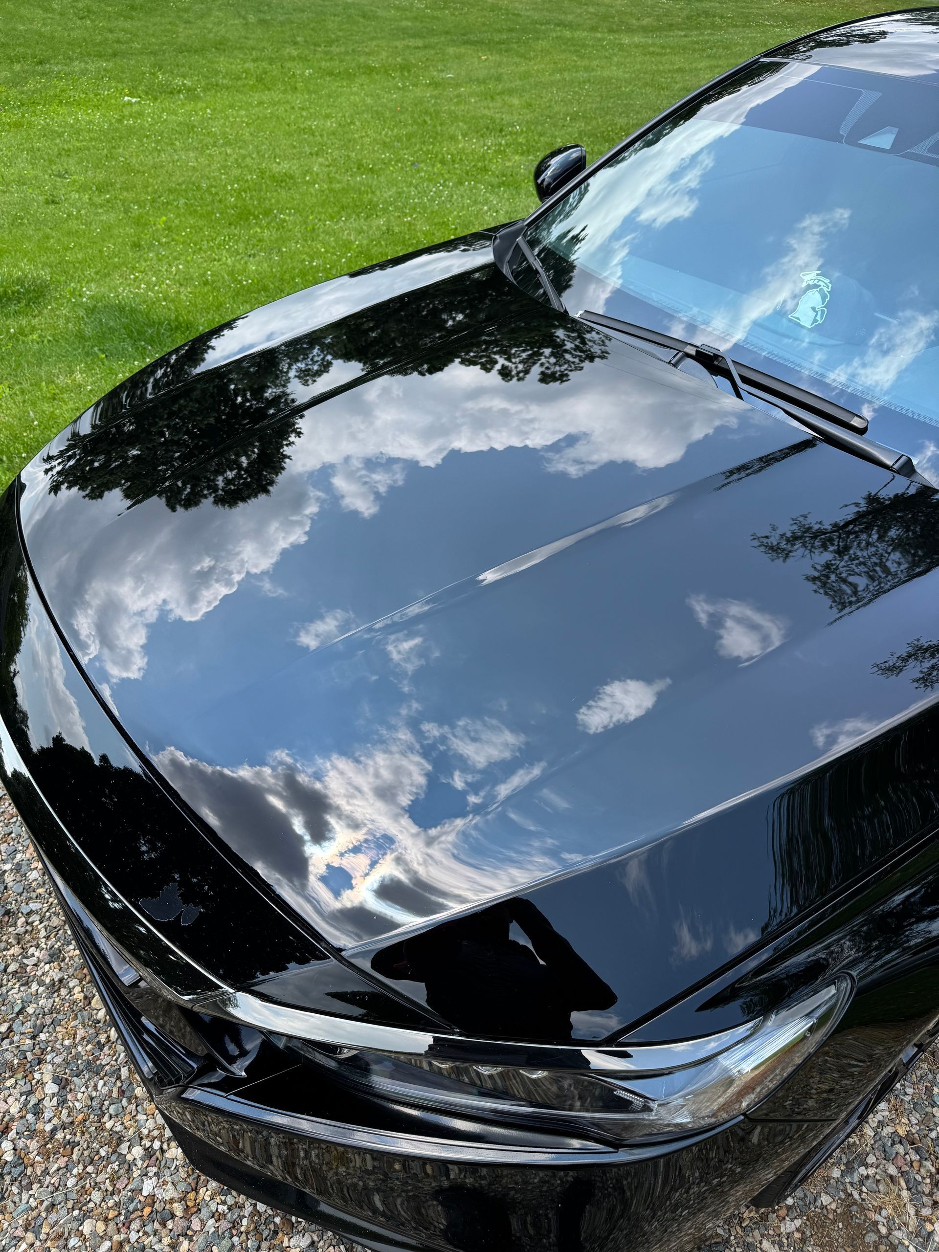 Black car hood reflecting a cloudy sky, sitting on a gravel driveway next to green grass.