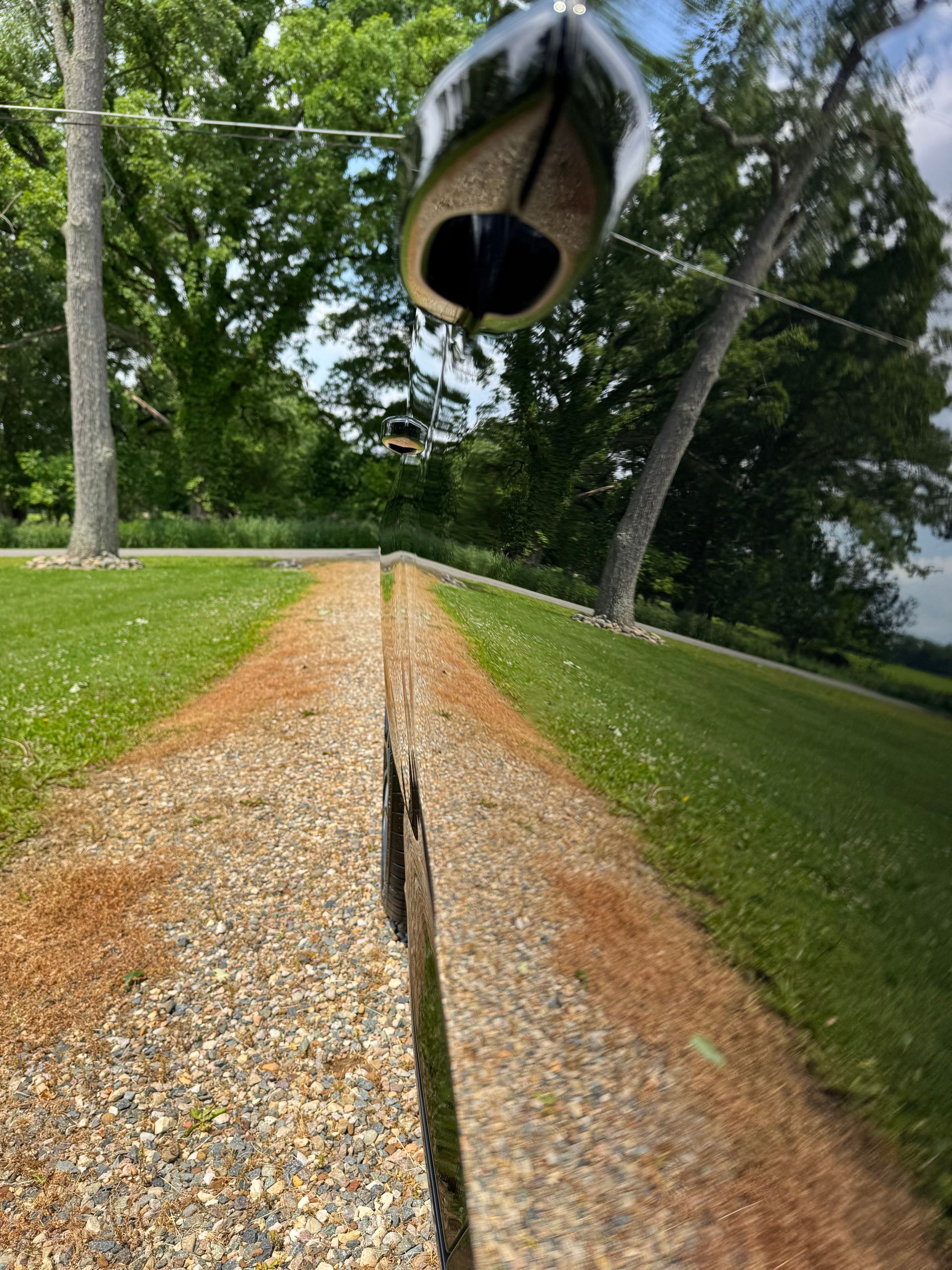 Gravel path flanked by green grass and trees, a shoe hanging overhead, reflected in a car door.