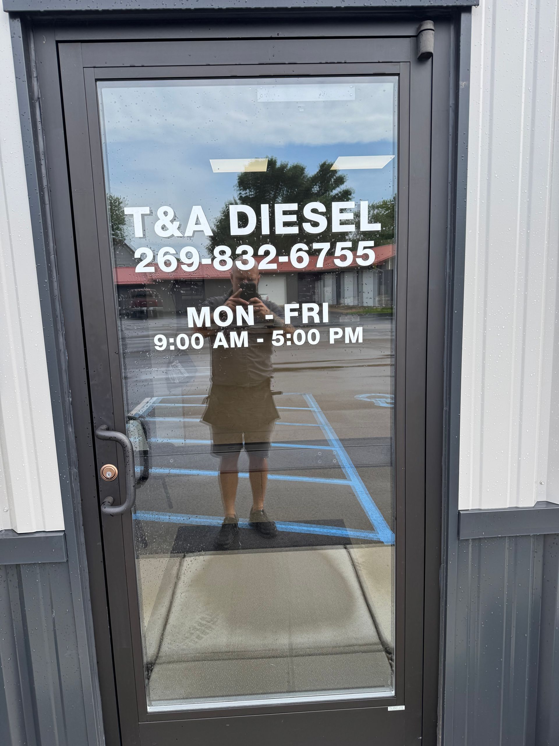Exterior door of T&A Diesel, with hours of operation. Person taking a photo reflected in the glass.