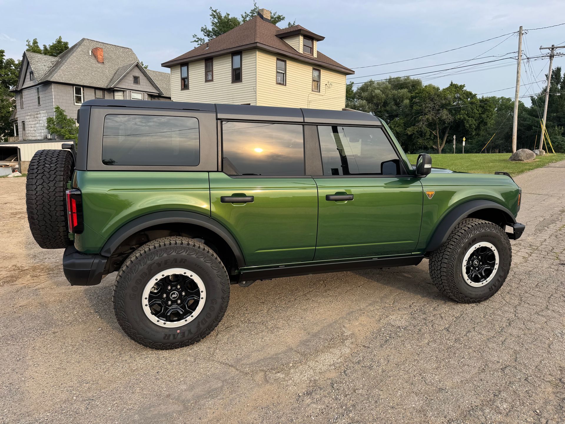 Green Ford Bronco SUV parked on gravel road with black accents; houses in background.