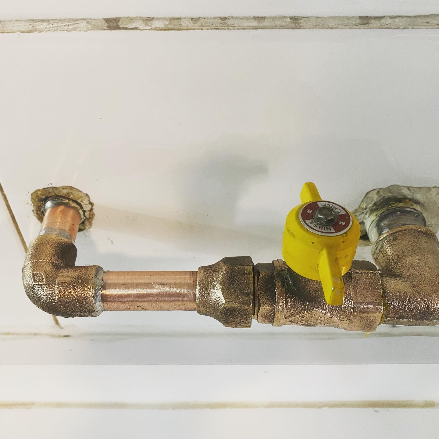 Tuberías y accesorios de cobre con una válvula amarilla sobre una pared blanca.