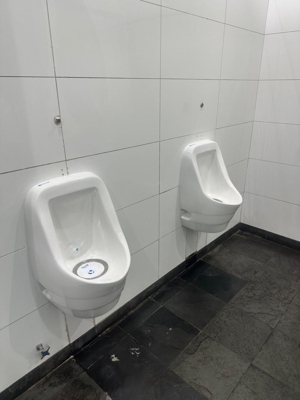 Dos urinarios blancos montados en una pared de azulejos blancos en un baño con piso oscuro.