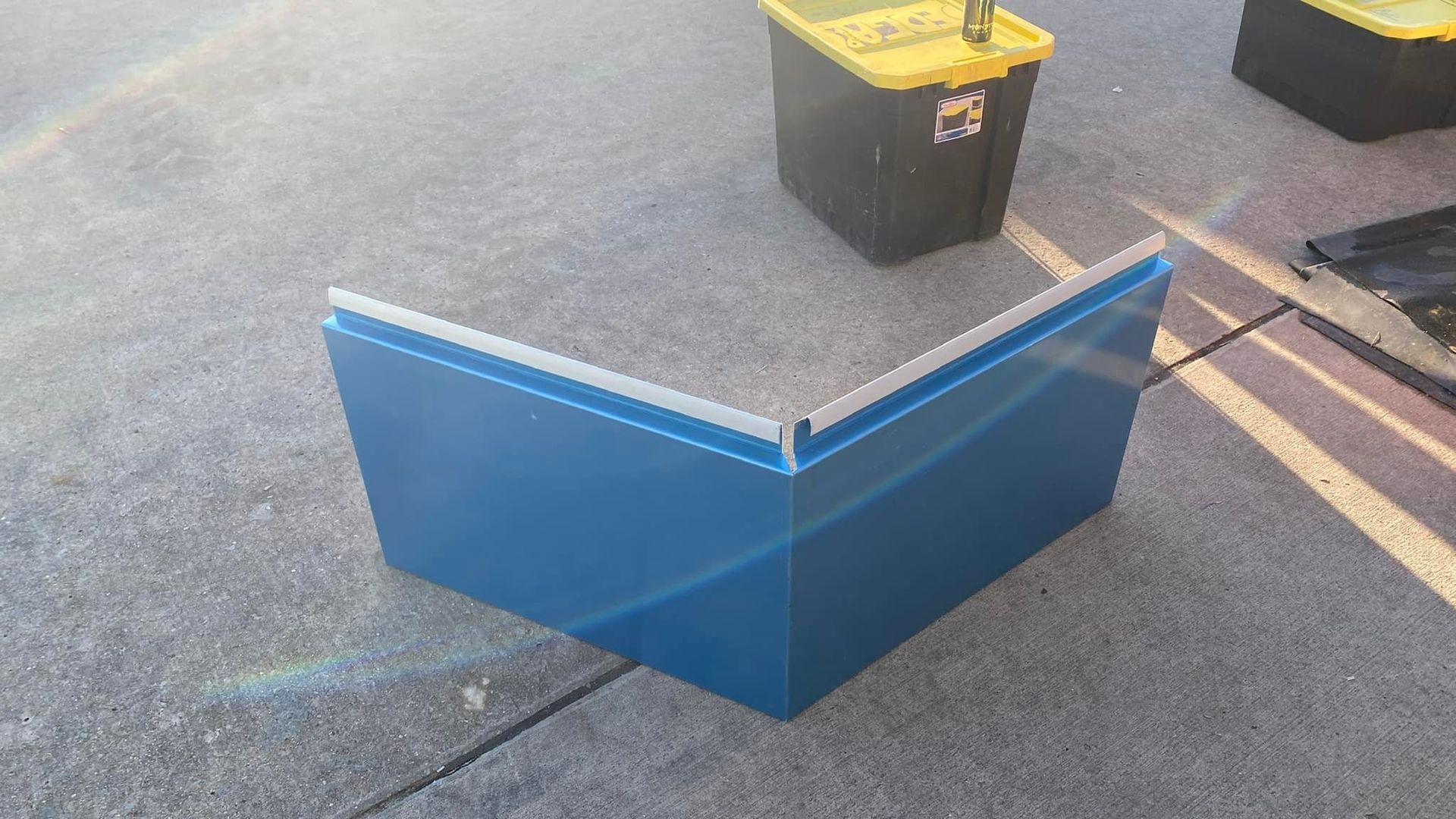 Blue metal sheet bent at a 90-degree angle, sitting on concrete, with storage bins in the background.