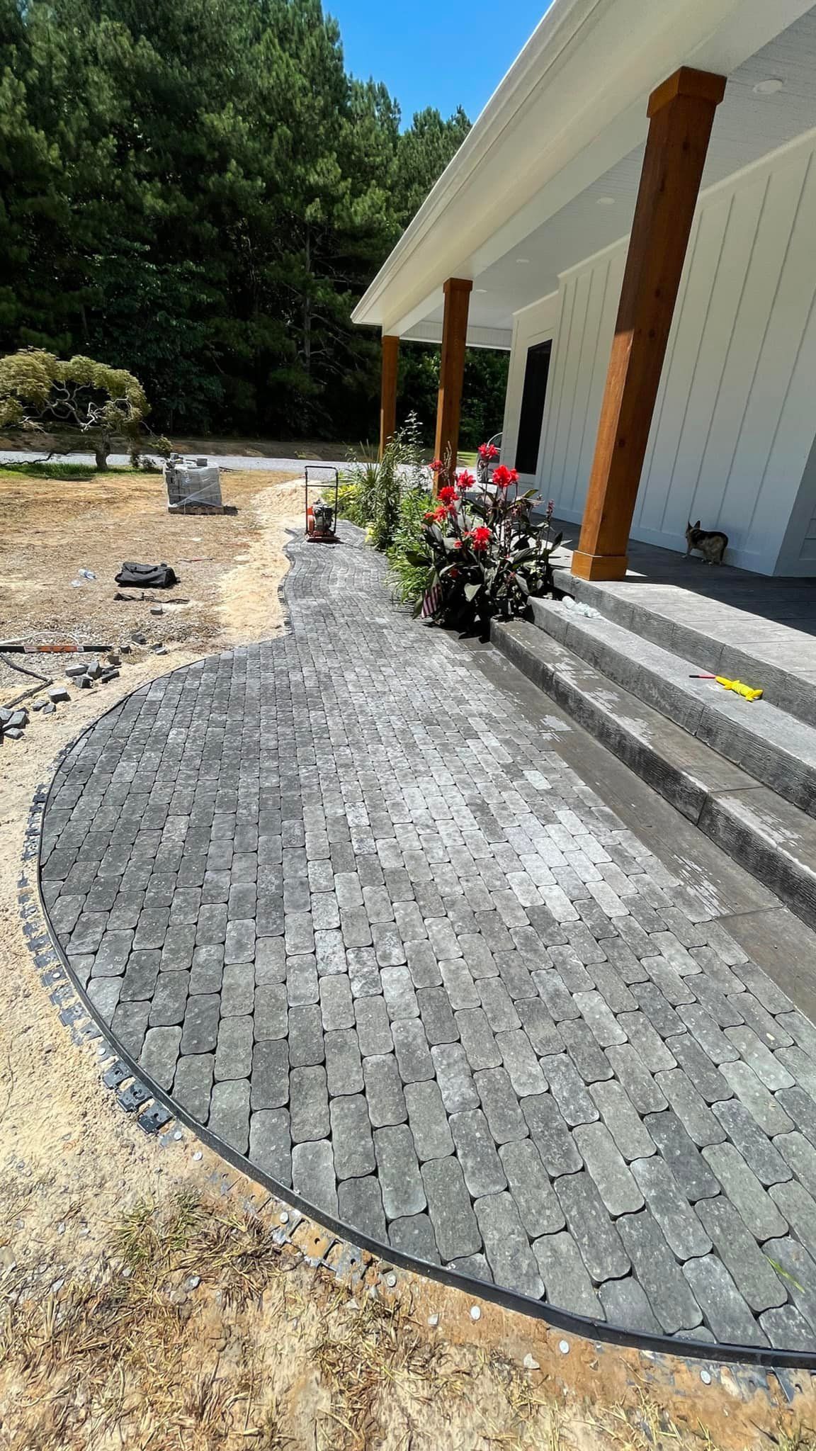 Gray brick pathway curves toward a white porch with brown columns, steps, and red flowers.