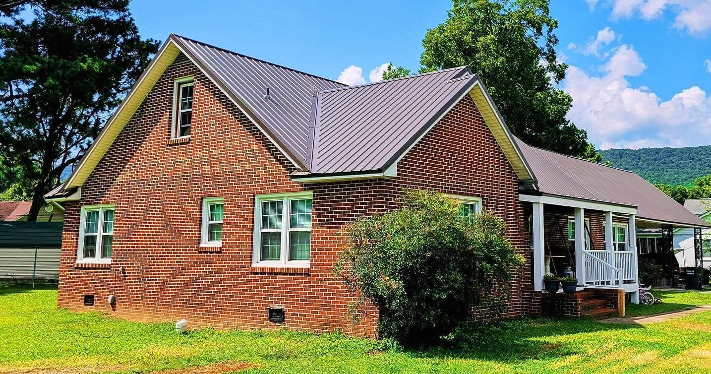 Brick house with metal roof, porch, and yard; mountains in background.