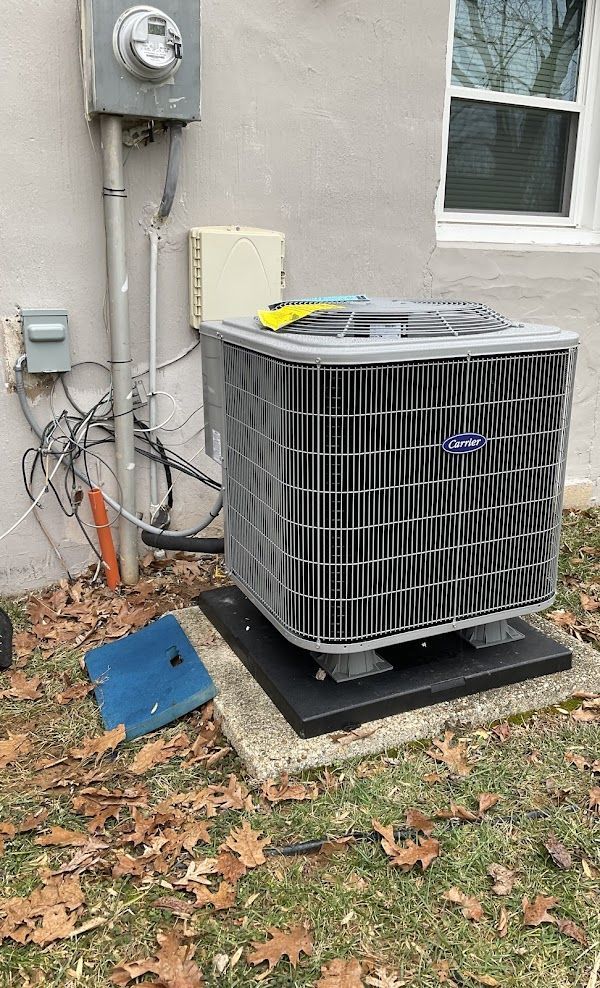Alex HVAC Service LLC | An outdoor air conditioner unit against a beige wall, with electrical components and fallen leaves.