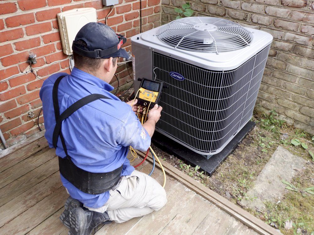 Alex HVAC Service LLC | HVAC technician kneeling, checking air conditioner unit outside on a brick wall.