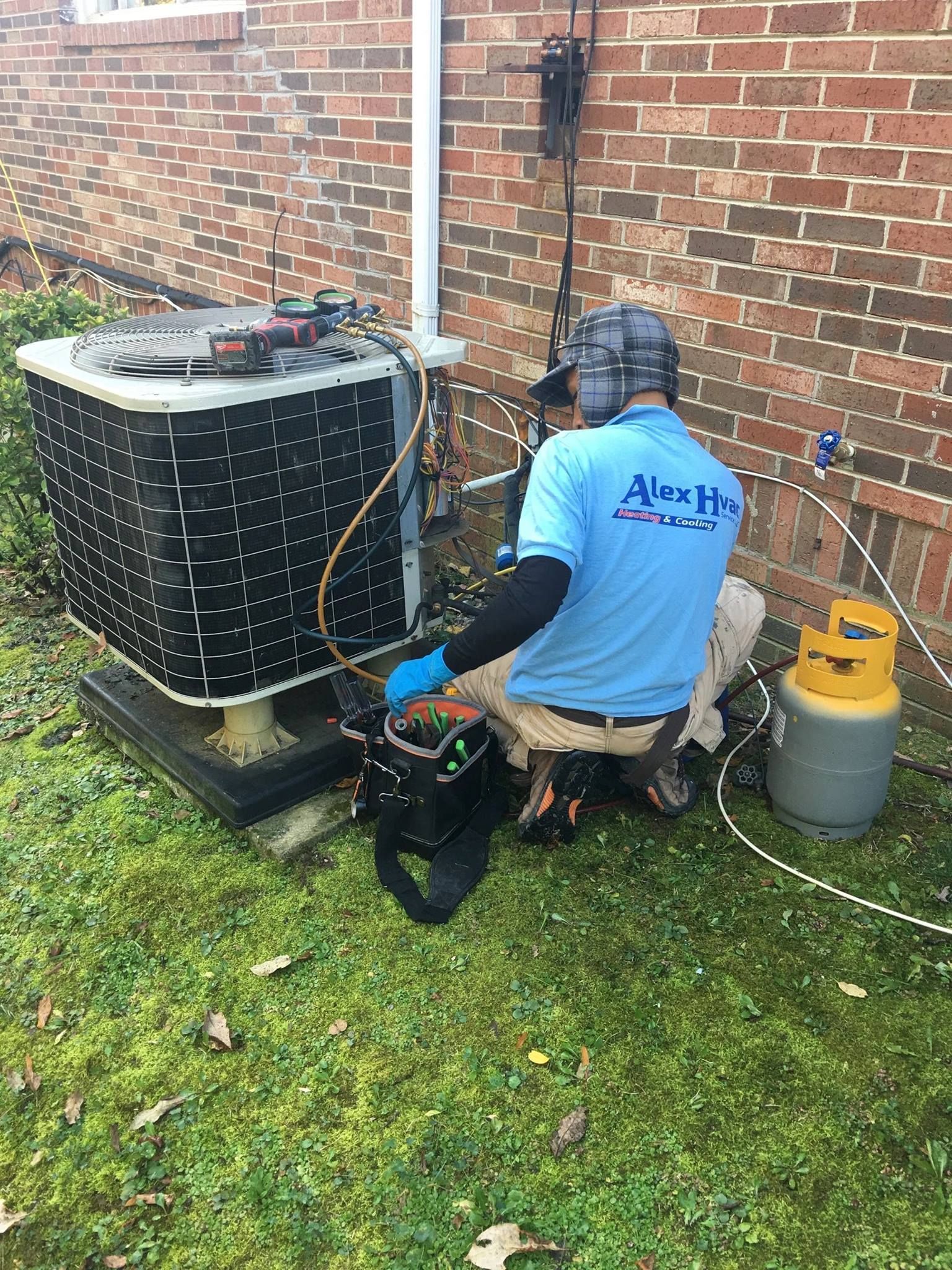 Alex HVAC Service LLC | HVAC technician working on an AC unit outdoors. He's wearing safety gear and has tools nearby.