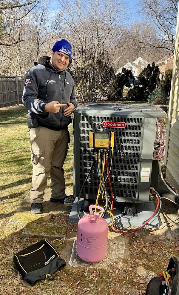 Alex HVAC Service LLC | An outdoor air conditioner unit against a beige wall, with electrical components and fallen leaves.