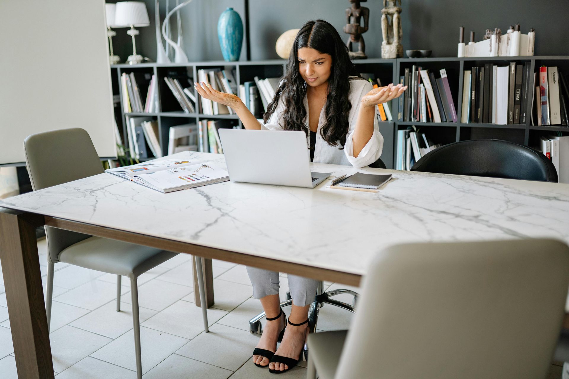 Woman with arms outstretched, looking confused at a laptop in an office setting.