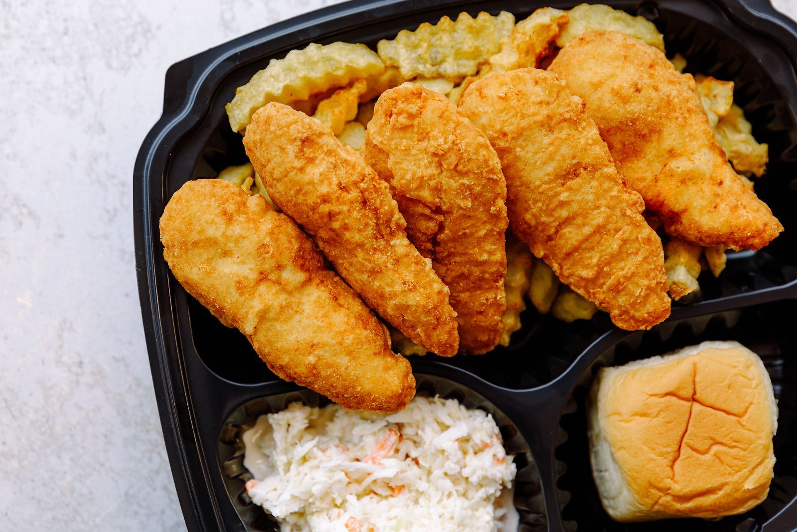 A black plastic container filled with fried chicken , coleslaw and french fries.