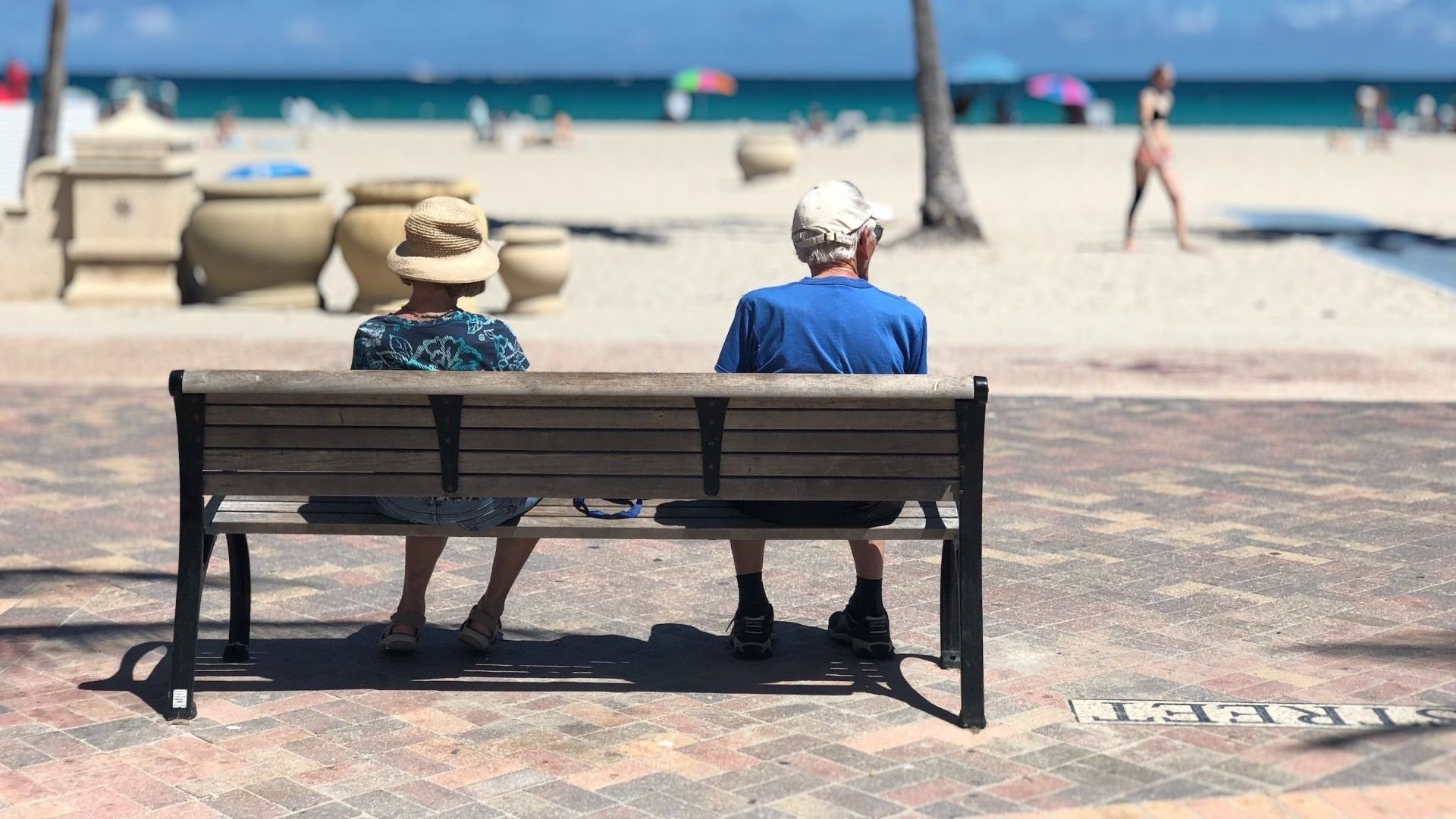 Two elderly people sit on a bench, facing the beach. The sky is blue, and people are enjoying the sun.