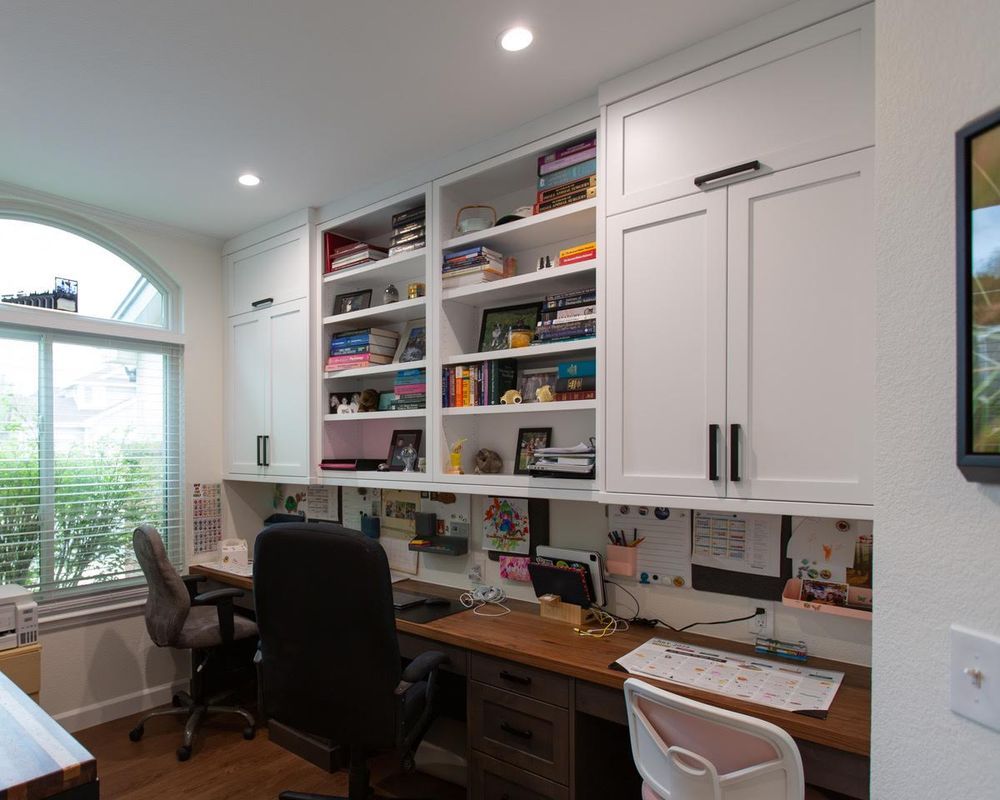 A home office with a desk , chairs , shelves and a window.