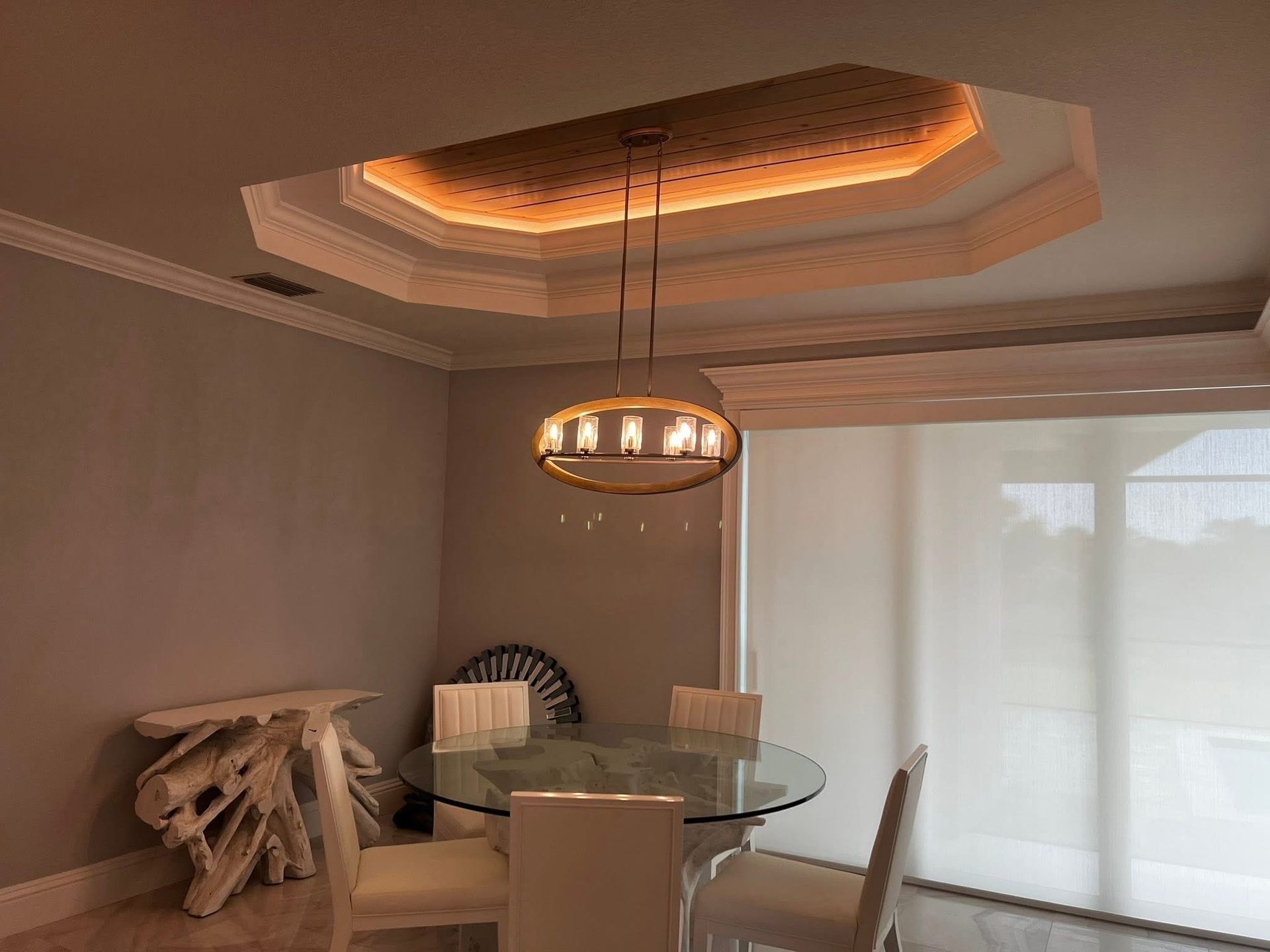 A dining room with a glass table, four upholstered chairs, a modern oval chandelier, and a recessed ceiling with lights.