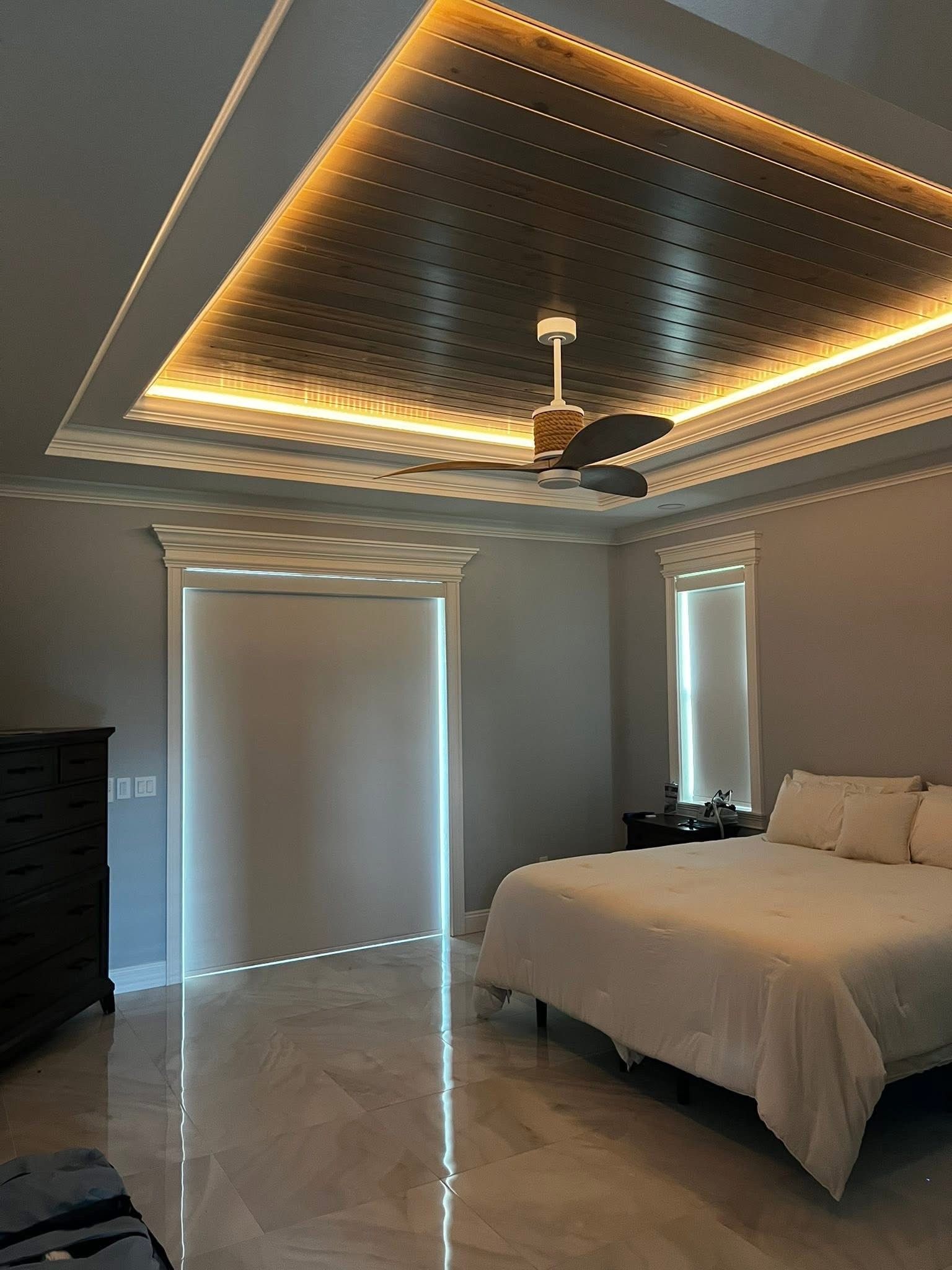 A modern bedroom with a tray ceiling featuring decorative metallic paneling, integrated warm LED lighting, and a fan.