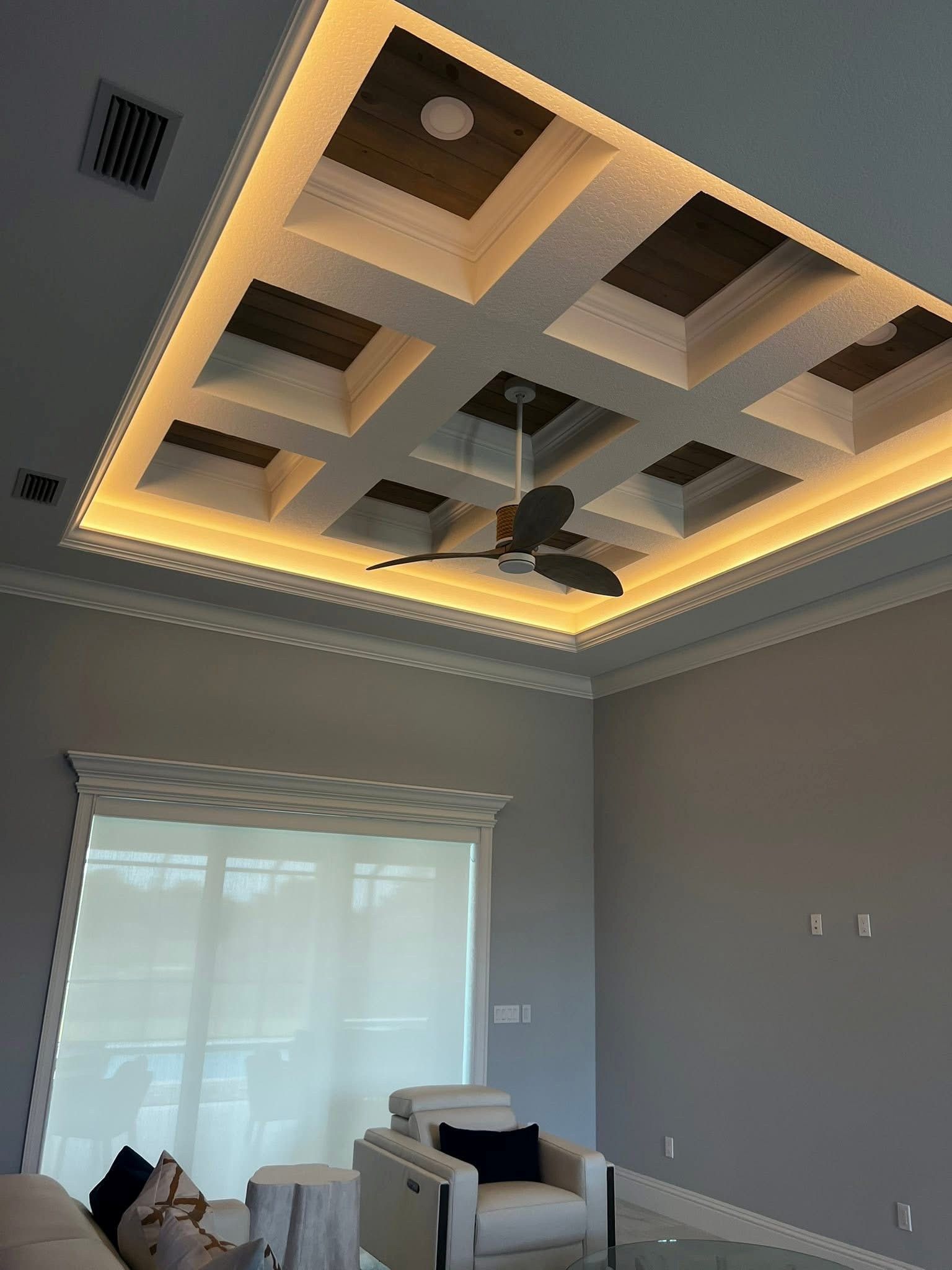 A low-angle view of a grey living room with a coffered ceiling, warm recessed lighting, and a dark wood ceiling fan.