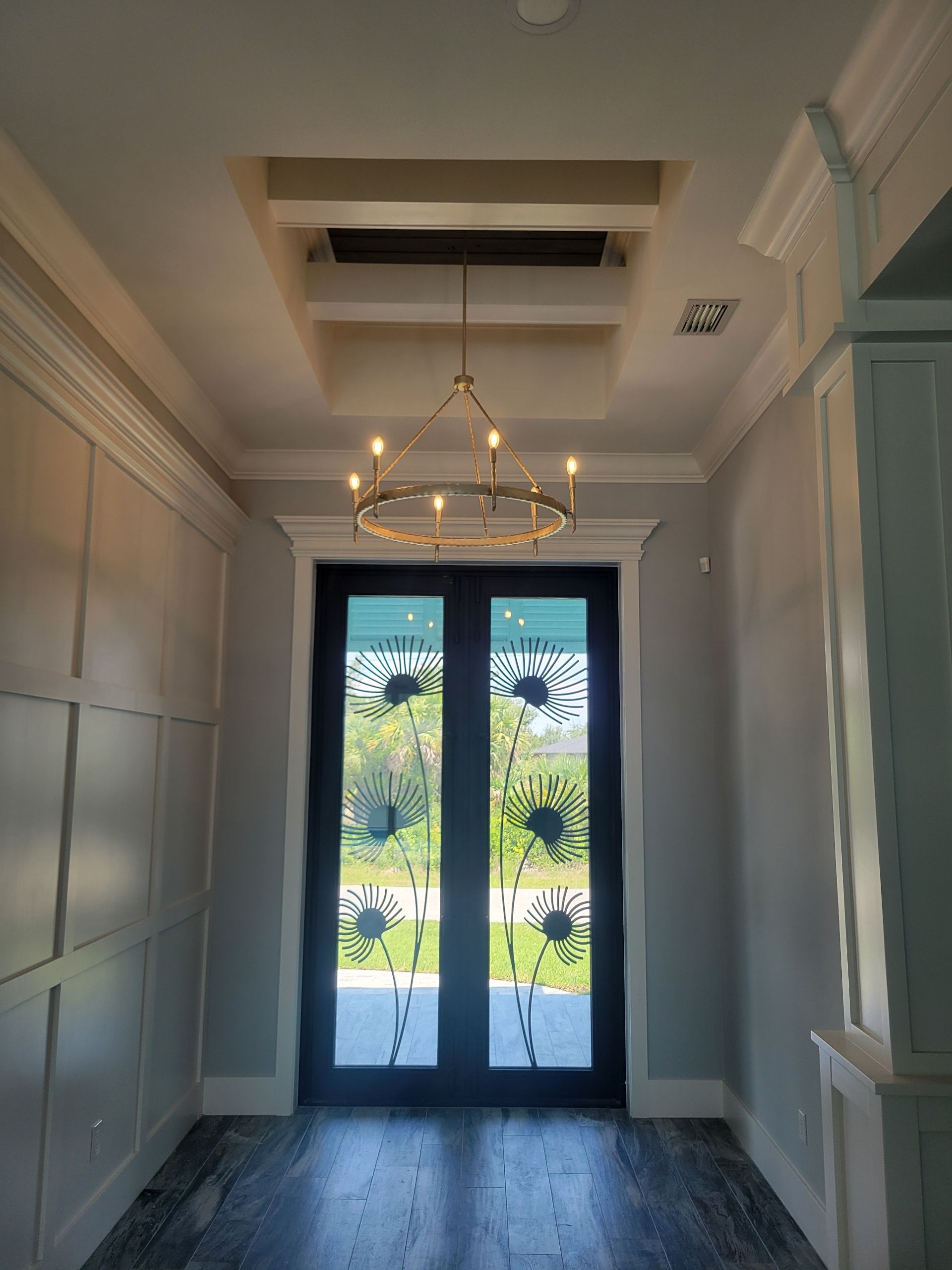 An elegant entryway with a dark double-door entrance featuring decorative glass, a brass chandelier, and light gray walls.