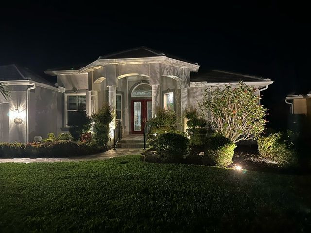 A suburban single-story house at night, illuminated by warm outdoor landscape lighting, featuring a red front door.