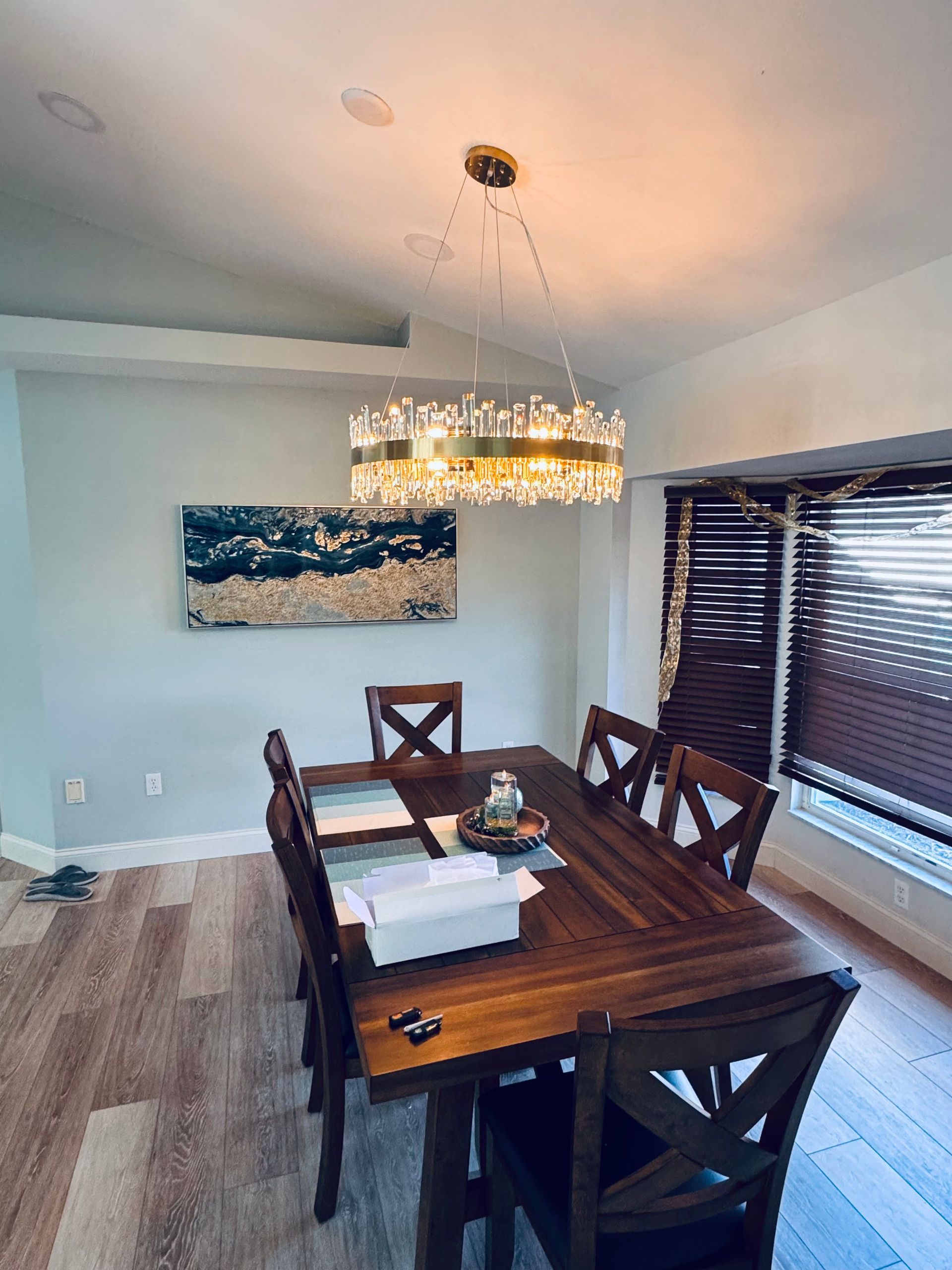 A dining room featuring a wooden table with cross-back chairs, lit by a crystal chandelier beneath a modern wall painting.