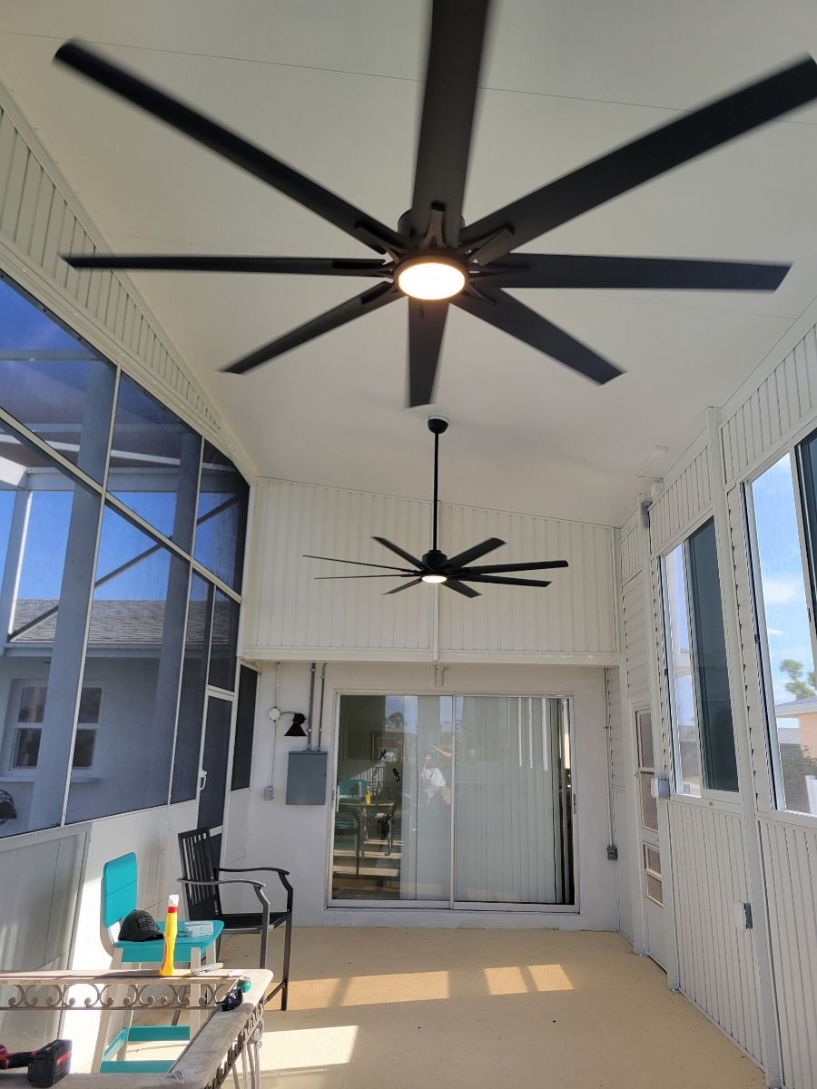 Two large black ceiling fans with lights hanging in a screened-in sunroom with white walls and a view of a house exterior.
