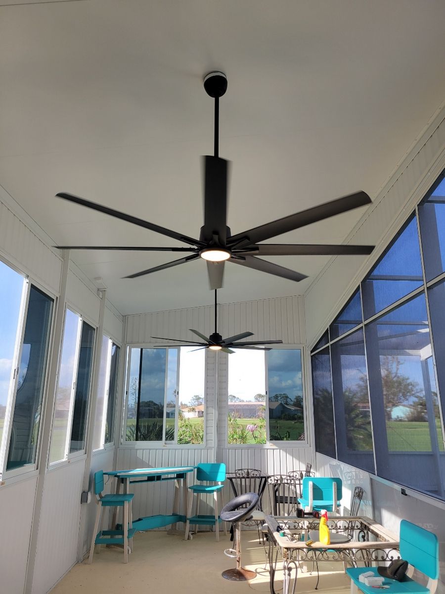 A sunroom with a large black ceiling fan, turquoise chairs, a small table, and glass-enclosed walls overlooking a field.