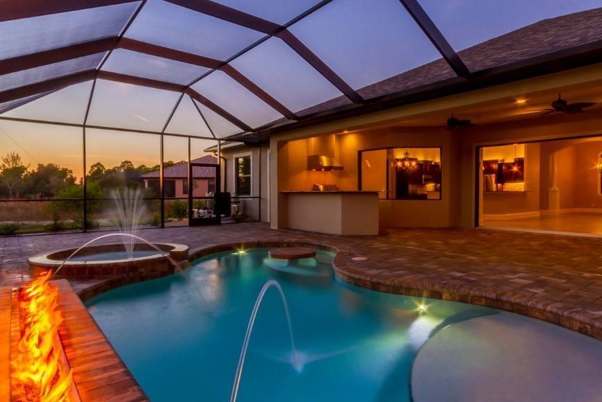 A backyard pool and hot tub enclosure at sunset, featuring a glowing fire pit, stone patio, and outdoor kitchen area.