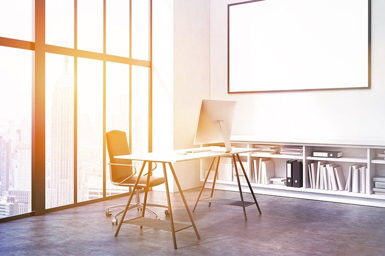 Bright office interior with large window, desk, chair, computer, bookshelf, and blank framed poster.