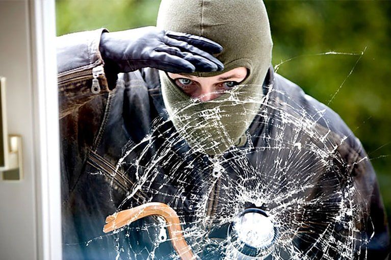 Person in balaclava peering through a shattered window, holding a crowbar.