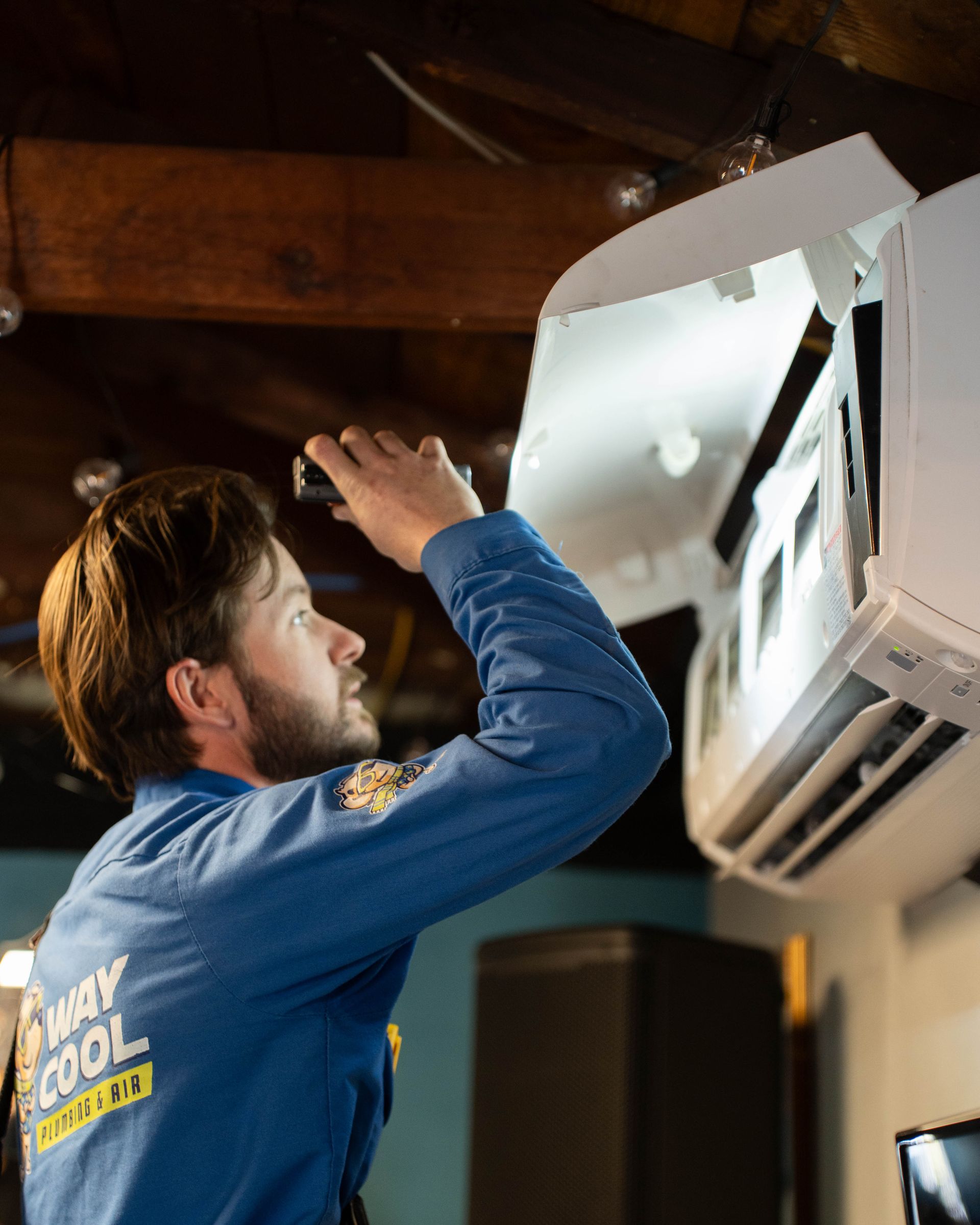HVAC technician in a blue Way Cool Plumbing & Air uniform inspecting the interior of a wall-mounted mini-split air conditioning unit, using a flashlight to check components during maintenance.