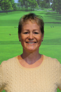 A woman in a yellow sweater is smiling in front of a golf course.